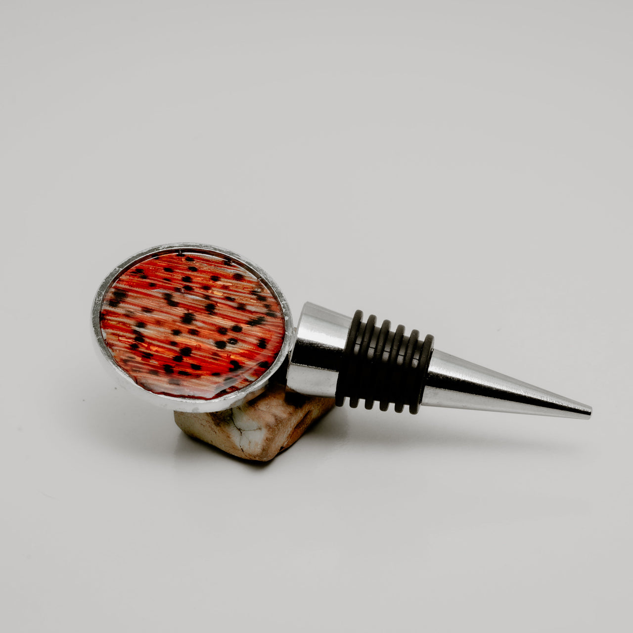 Silver wine bottle stopper with round lionfish tail fin resin charm in red and black pattern displayed on natural stone on white background