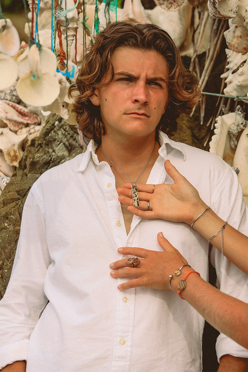 A man in a white shirt wearing a silver bar necklace in front of hanging shells. Two hands in front of his shirt show four bracelets and three rings and one is holding his necklace . 