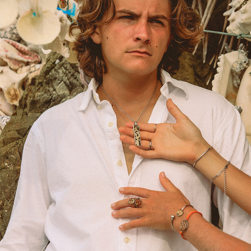 A man in a white shirt wearing a silver bar necklace in front of hanging shells. Two hands in front of his shirt show four bracelets and three rings and one is holding his necklace . 