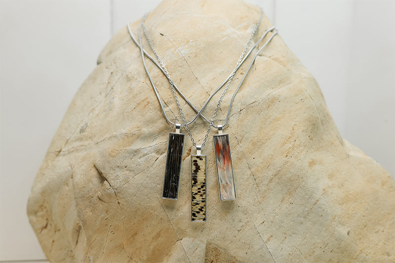 Three silver lionfish fin bar necklaces with tail, pectoral, and pelvic fin resin pendants in black, speckled, and amber colorways displayed on natural stone