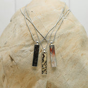 Three silver lionfish fin bar necklaces with tail, pectoral, and pelvic fin resin pendants in black, speckled, and amber colorways displayed on natural stone