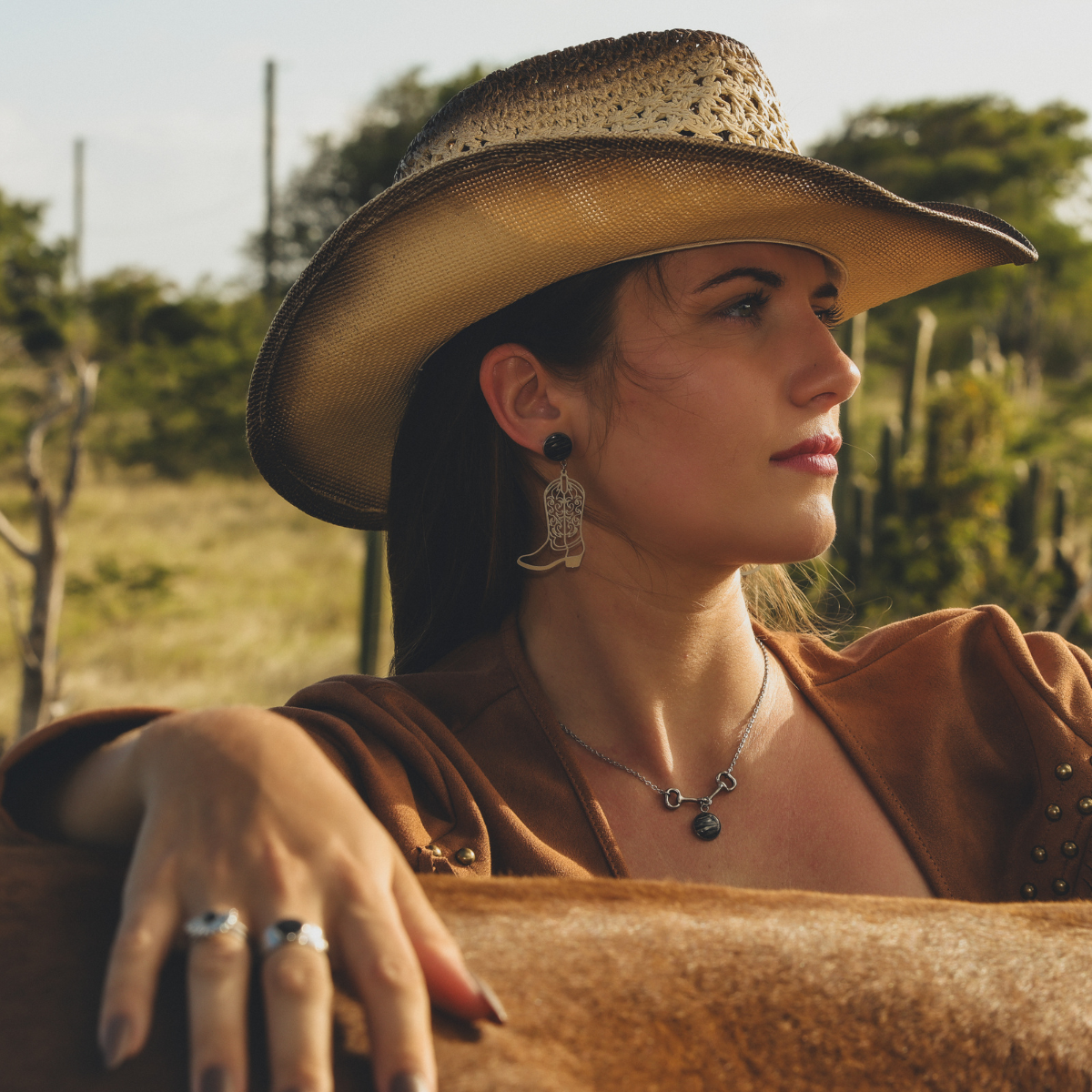 Woman in cowboy hat wearing lionfish jewelry with boot earrings, silver necklace, and rings outdoors